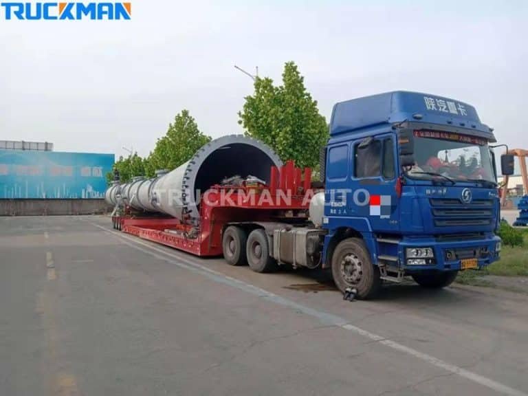Blue heavy-duty truck with low-bed semi-trailer carrying oversized cylindrical industrial equipment on city roads.